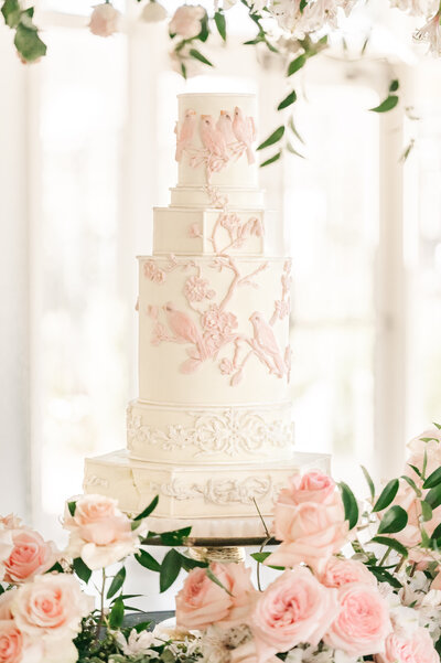 A white wedding cake with pink piping surrounded by pink and white flowers and greenery at Donovan Manor by JoLynn Photography, a destination wedding photographer