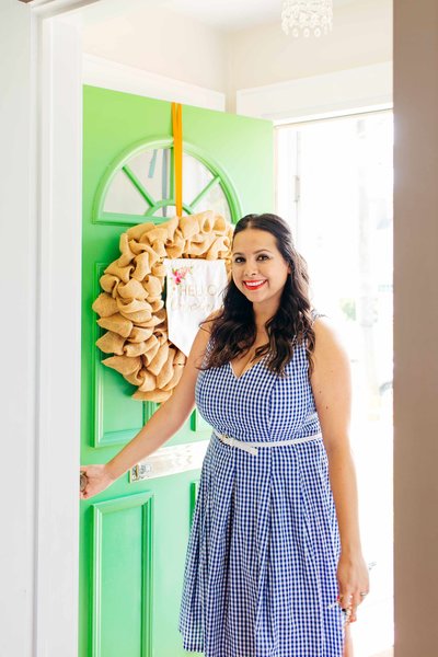 A woman in a blue gingham dress holding open a bright green door.