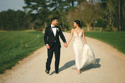 A wedding couple holding hands and walking along a dirt path.