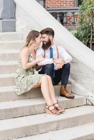 Engaged couple sit on steps and smile at each other forehead to forehead during engagement photography session