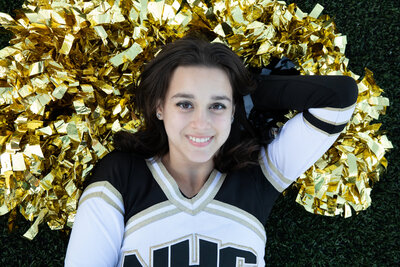 A teenage girl in a black and white cheerleading uniform lays on gold pom poms Nanuet senior portrait photographer Milestone Images