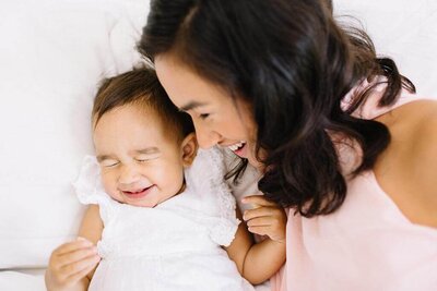 Baby girl in a white dress with mommy lying down giggling together.