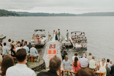 Couple getting married on their parents' dock with wedding party sitting in boats