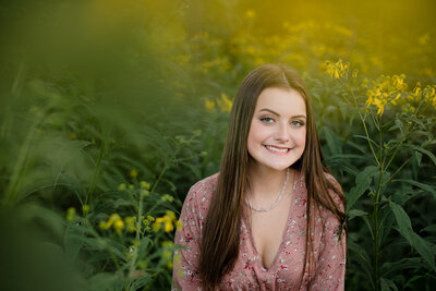 teenage girl with long straight brown hair sitting in tall yellow flowers with yellow bokeh