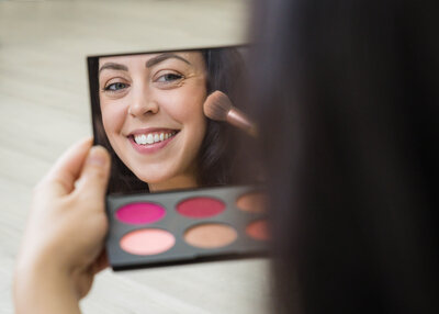 woman holding a makeup palette and smiling into the mirror