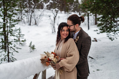 A couple stands on a snowy bridge in a winter forest in Tromsø, Norway. The woman, wearing a beige cardigan over her wedding dress and holding a rustic bouquet, shares a warm smile with the camera, while the man, in a patterned brown suit, gently kisses her on the forehead, a picture of affection and coziness amidst the cold on their elopement day.