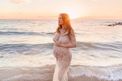 pregnant woman in lace dress stands next to the water on the beach for stunning photos during her Wailea maternity photoshoot at sunset