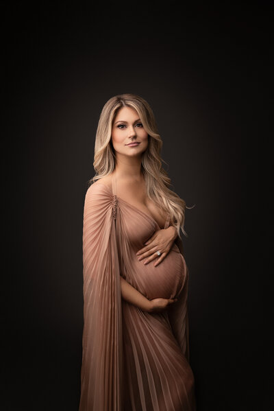 Woman poses for a fine art maternity photoshoot. She is wearing a dusty rose couture gown with that has a ruched floor-length caped sleeve. She is standing angled to the camera with one hand above and one hand below her baby bump. She has a closed-mouth smile and her hair is draped over her left shoulder. Captured by best Main Line Philadelphia maternity photographer Katie Marshall.