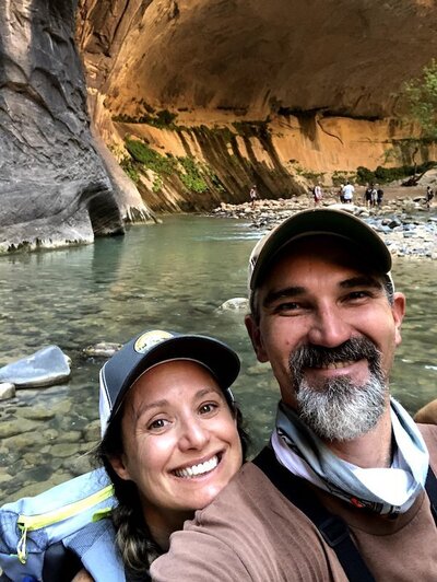 Alicia and Errin, co-founders of EnForma, smiling during a scenic hike through the Narrows in Zion National Park, reflecting their adventurous spirit and commitment to active, nature-based wellness.