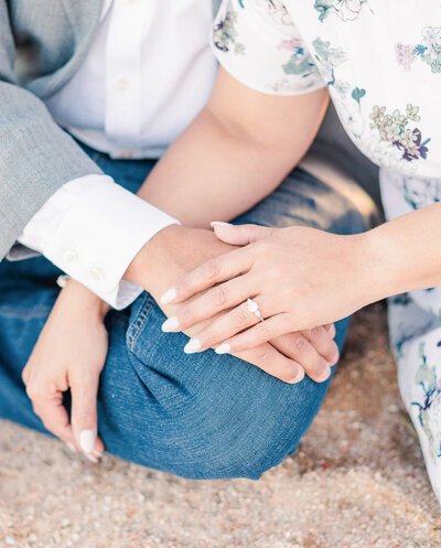 A couple sits on a beach holding hands during their engagement session.