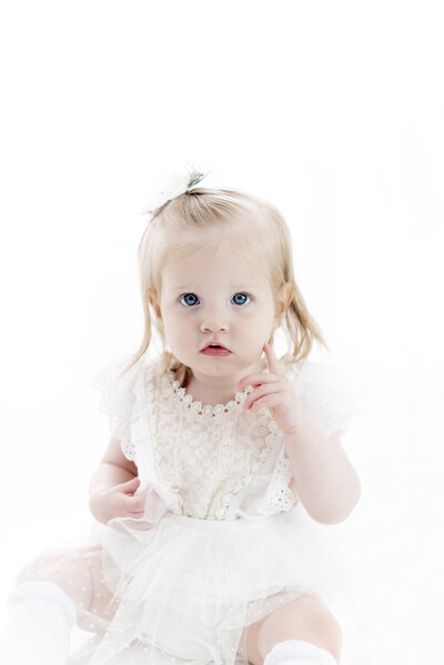 A toddler girl holds up one finger while sitting in a studio in a white lace dress as posed by a childrens photographer Atlanta