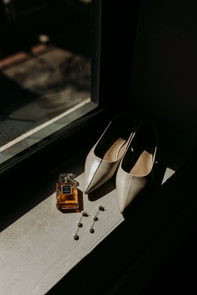 Bridal shoes and perfume bottle and earrings sit on window sill in contrast stark lighting