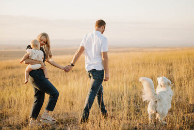 Husband walking and holding his wife's hand while the wife is holding their baby and their dog is walking alongside them