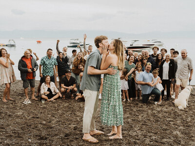 A couple shares a kiss at the West Shore Cafe in Tahoe, as a crowd of guests celebrates around them. The guests, dressed in casual summer attire, are cheering and raising their arms in the air. The scene takes place on a sandy shore with boats visible on the water under a cloudy sky, capturing the essence of a special occasion at this lakeside venue. The photographer captures this candid moment, conveying the joy and excitement of the event.
