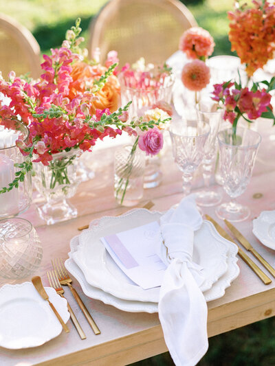 Wedding reception table close up: warm orange, pink and red flowers, scalopped white dishes, and tied white handkerchiefs