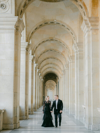 a couple in black outfits walking towards the  camera while smiling and laughing at one another in a tunnel of arches at the Louvre