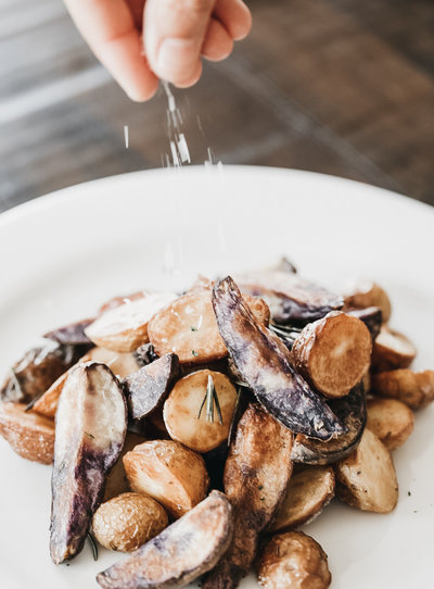 Photo of a plate of rosemary potatoes being seasoned by the fingers of the chef. Salt is sprinkled on top and the grains are blurry with motion. The potatoes are on a white plate and wooden table and look delicious.