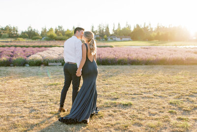 A photograph from behind as engaged couple kiss in a lavender field wearing cocktail attire