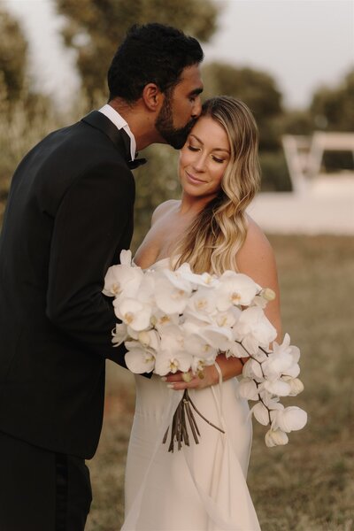 Bride and groom kissing each other. Bride holds an orchid bouquet. They stand in the olive grove of Masseria Moroseta, Ostuni