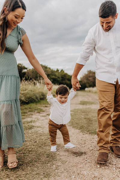 A family of three walking their toddler down a San Antonio trail. Mom holds one hand, dad holds another hand, and the toddler walks in the middle.