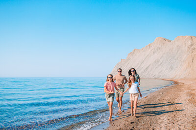 A family of four, two adults and two children, joyfully walk along a sandy beach by clear blue water. Rugged cliffs rise in the background under a vibrant blue sky. They are dressed in casual summer clothes, smiling and enjoying the sunny day.