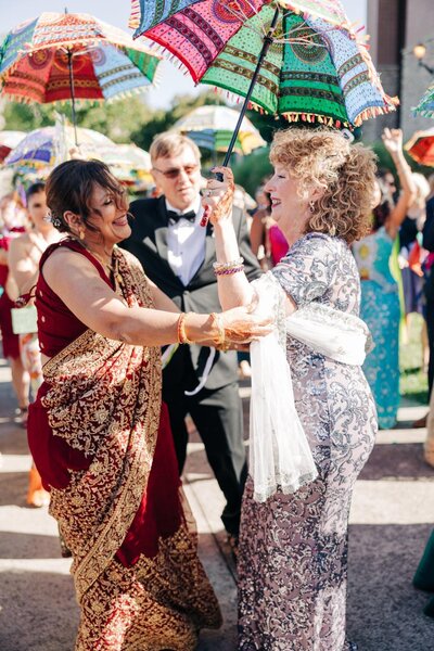 Two women in ornate dresses dancing with colorful umbrellas at an outdoor celebration.