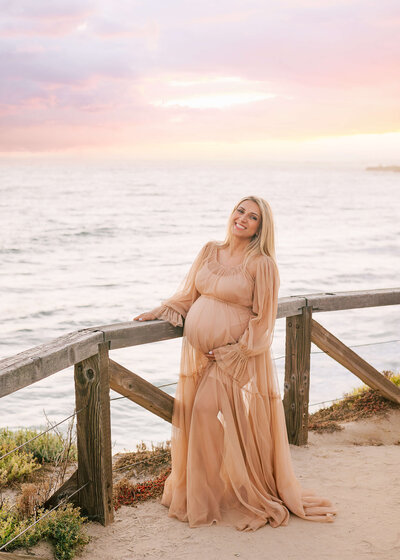 Mama standing above ocean in Newport Beach smiling and holding baby bump by Ashley Nicole.