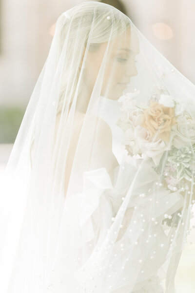 Bride poses for a wedding day portrait. Her veil is over her face and her bouquet under the veil. She is looking down at the flowers. Image is very light and airy. Captured by best North Shore Boston Wedding Photographer Lia Rose Weddings