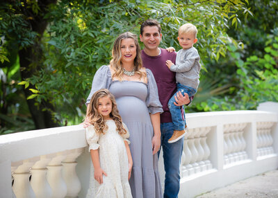 Family portrait featuring a mother in a light purple dress, a father holding a young boy, and a young girl in a white dress, all smiling and standing outdoors next to a white decorative railing with greenery in the background.