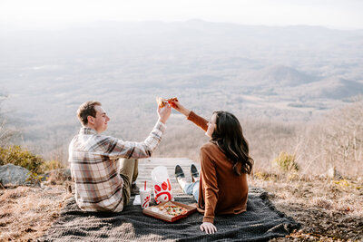 man and woman clinking glasses while sitting on ground with chick fil a picnic