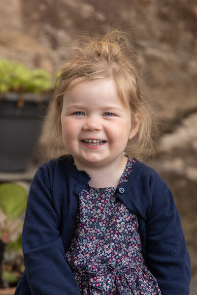 Jente på tre år iført marineblå cardigan og lilla blomstrete kjole smiler mot kamera.