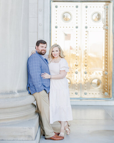 A guy leans in his blue blazer and khaki pants against a column with his girl in a light flowy dress in front of the capitol