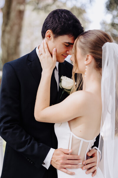 Bride and groom stand forehead to forehead, as the bride gently rests her hand on the groom's face. The bride wears pearl earrings and the groom has a white rose boutonniere. Photo taken by Orlando Wedding Photographer Four Loves Photos and Film.