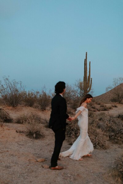 A bride and groom in wedding attire walk through the Phoenix, Arizona desert during their elopement.