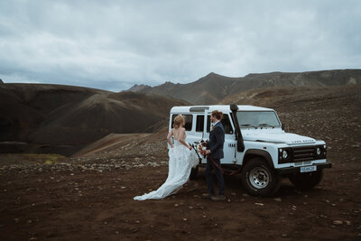 the bride and groom are walking towards their land rover that they used to drive through iceland. the groom is opening the car door for the bride, and the bride is holding her dress up as she gets in.