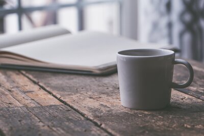 A mug sits on a table with on open book in the background, a thoughful  scene.