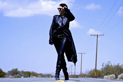 Portrait of Female Musician Leslie Cours Mather walking down road in long coat hand to sunglasses she wears telephone poles behind her