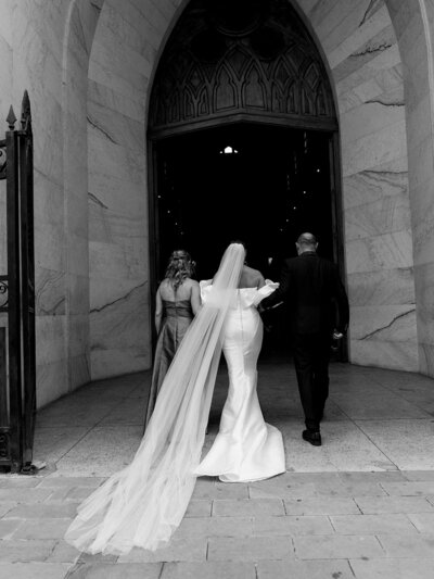 The bride entering the church in a very editorial style photograph. It is her back with her parents walking her in.