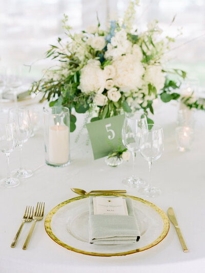 Reception place-setting with wine glasses, candles, gold cutlery, gold rimmed plate, and large white and green bouquet in the background