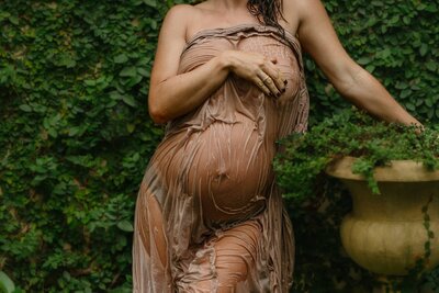 Close-up of a pregnant woman's belly wrapped in wet fabric during a maternity photography session, standing against lush green foliage.