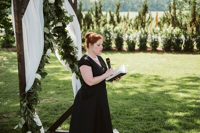 Raina van Setter, Nashville wedding officiant, dressed in black stands in front of an arbor covered in drapery and green garland with white flowers