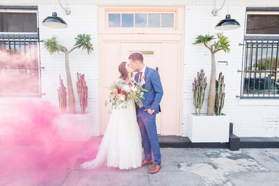 Bride and groom kiss on their wedding day holding beautiful bouquet as puff of pink smoke blows in from side