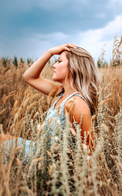 Senior girl runs hands through hair while sitting down in tall grass and sage weeds