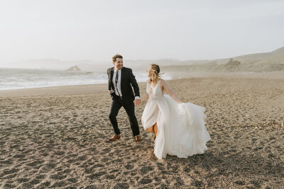bride and groom holding hands running on beach