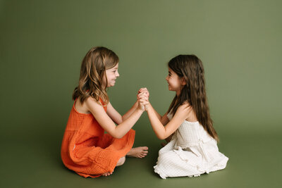 two girls sitting criss cross on the floor and holding hands playfully
