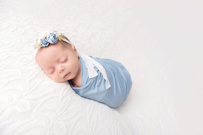 A newborn baby sleeps in a  blue swaddle and matching floral headband on a white bed