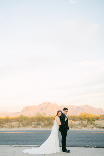 The bride embraces the groom, who stands with his back against the backdrop of roads and the stunning Superstition Manor mountains, capturing a moment of love against a scenic setting