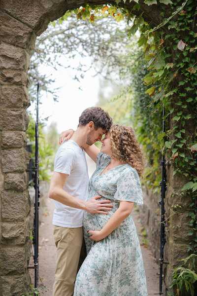 couple in pretty field with green trees poses holding baby bump during boston maternity session