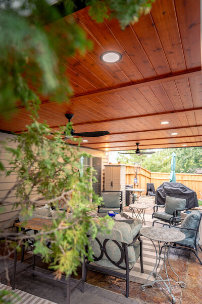 Looking through tree branches at a patio underneath a deck with a cedar ceiling and recessed lights. 