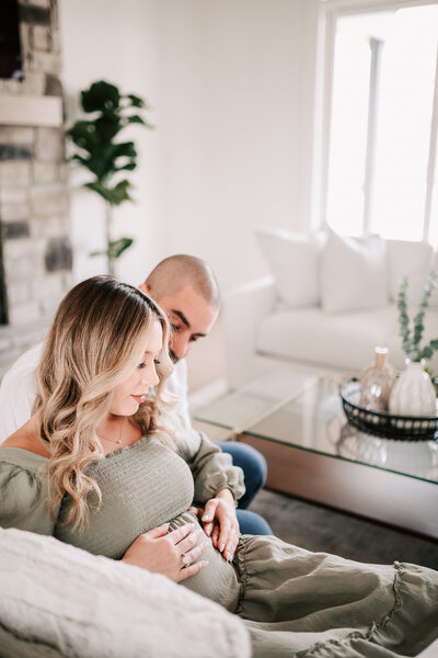 Pregnant new mom in a sage green dress sitting on an ivory couch in a brightly lit white room with her husband behind her both cradling her belly